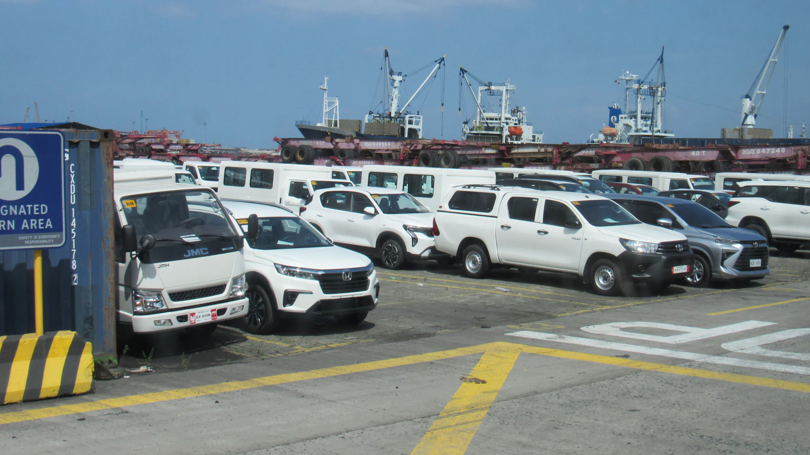 Picapes e carros novos estacionados em pátio portuário, com navios e guindastes ao fundo